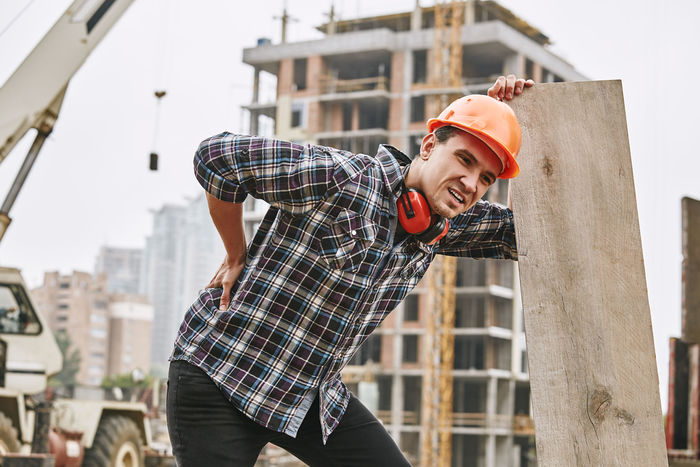 Hard work. Construction worker in protective helmet feeling back pain while working at construction site. Building construction. Pain concept. Dangerous job